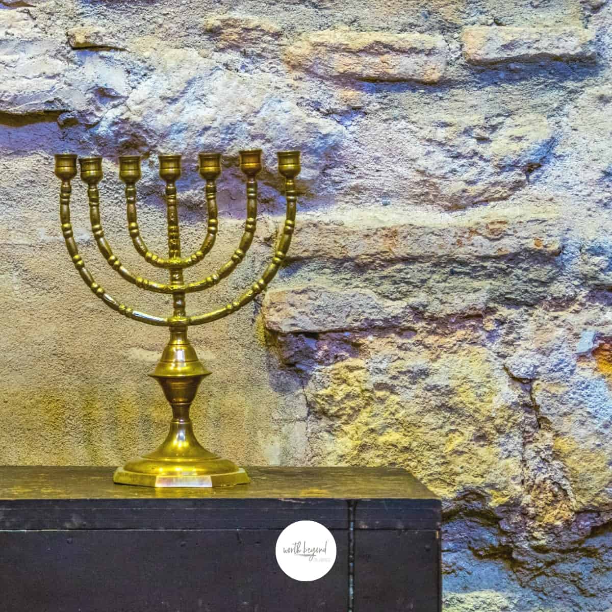 a menorah on a wooden table against a stone wall and the Worth Beyond Rubies logo at the bottom of the image