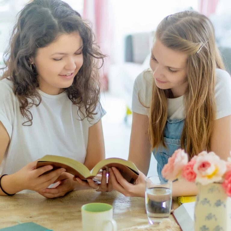 two teenage girls sitting together at a table, smiling and looking at a Bible