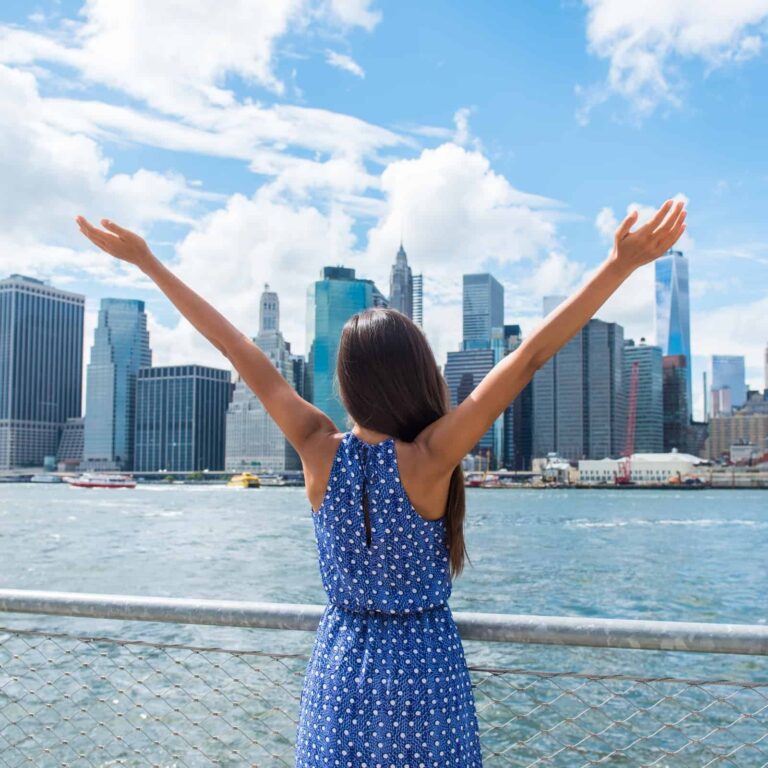 An image of a woman standing on the river facing the New York City skyline and she is wearing a blue, polka dot dress and her arms are up in the air toward the sky - Freedom in the Bible