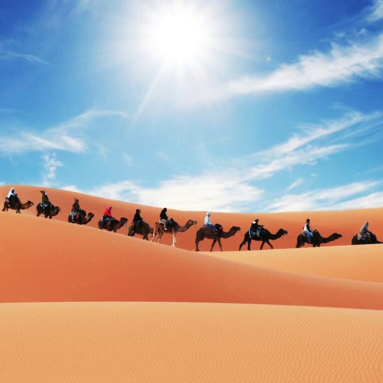 An image of a caravan of camels and riders in the desert against a bright blue sky