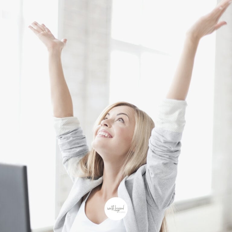 An image of a blonde woman in a gray suit and a white top sitting at a desk with her hands up in the air looking upward For It Is God Who Gives You Power to Get Wealth