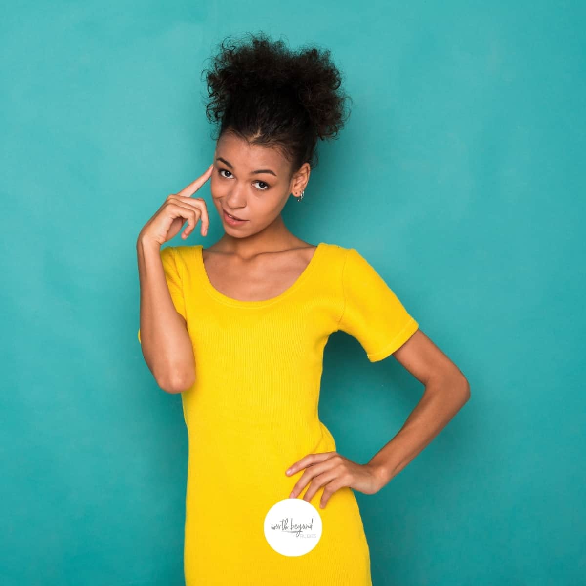 A beautiful black woman in a yellow dress standing in front of a blue background with her pointer finger up to her temple for post called The Mind is the Battleground - Protecting Against Spiritual Attacks