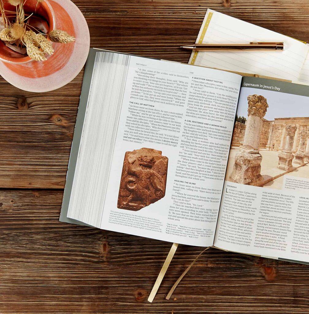 An image of the Holy Land Illustrated Bible, opened to a page on a wooden table with a plant next to it