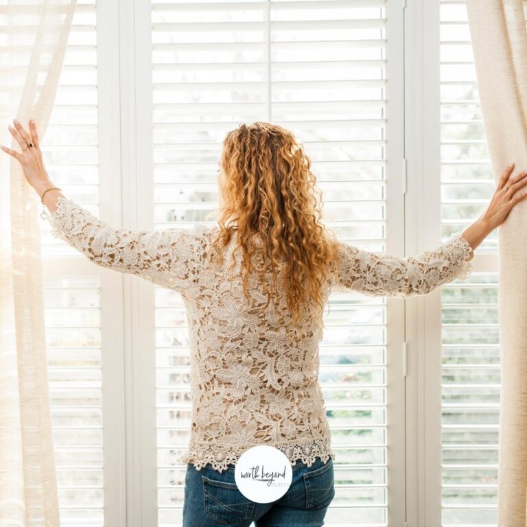A woman with long curly blonde hair facing a window drawing back curtains and the Worth Beyond Rubies logo on the bottom for post on Searching for God