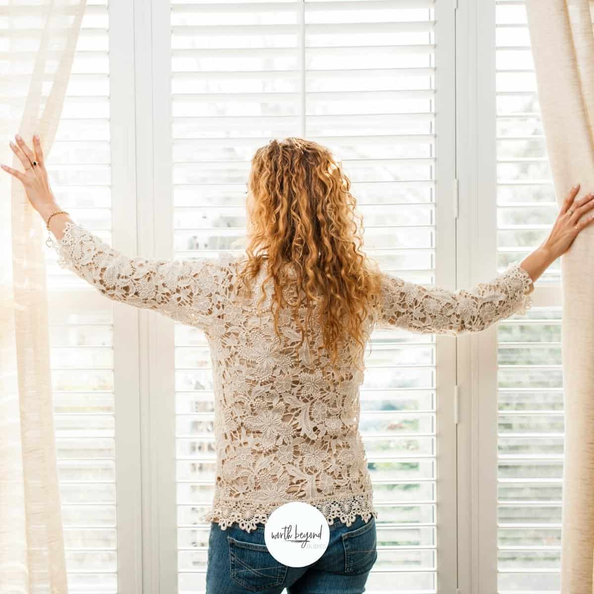 A woman with long curly blonde hair facing a window drawing back curtains and the Worth Beyond Rubies logo on the bottom for post on Searching for God