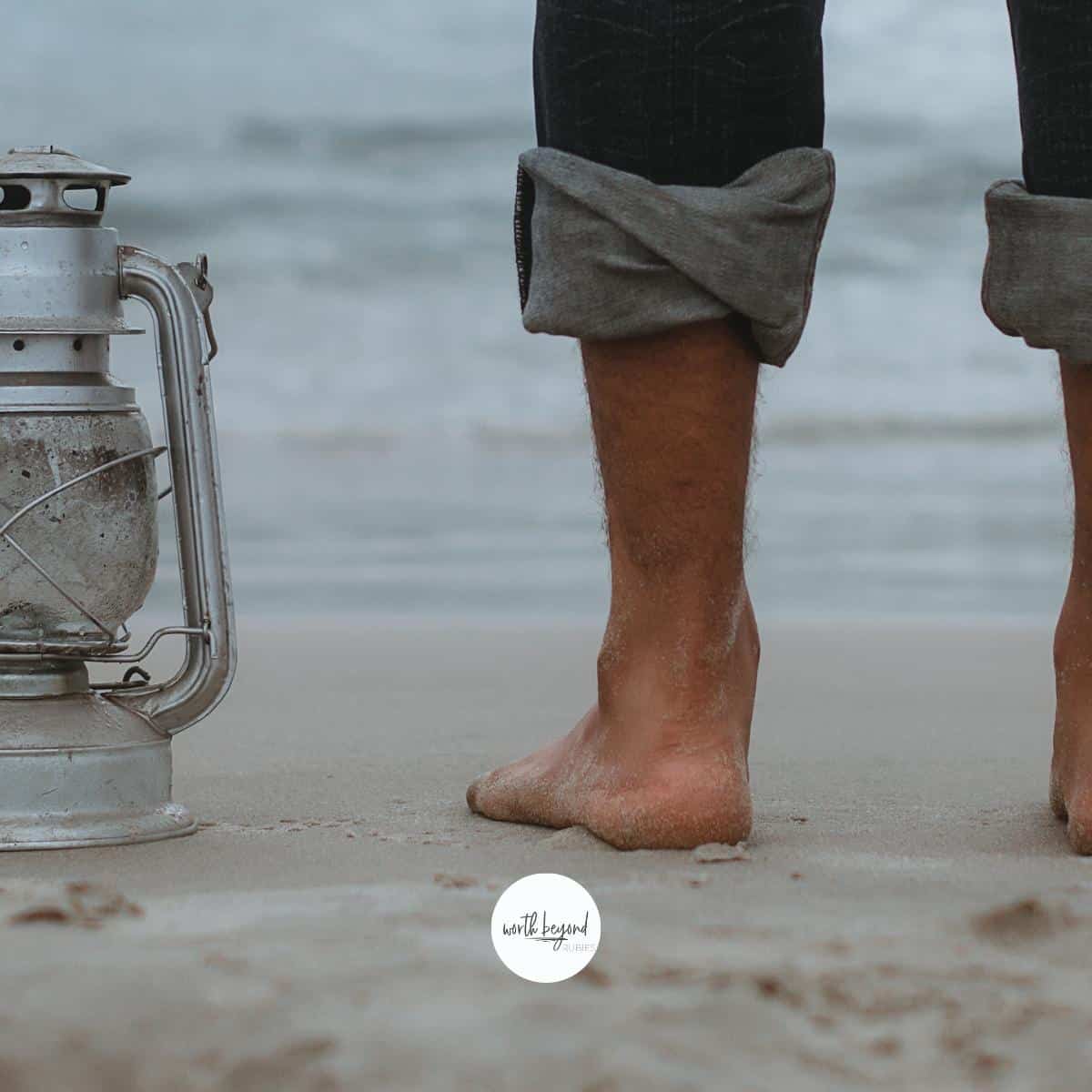 A man's legs with jeans rolled up, standing on a beach next to a lantern and the Worth Beyond Rubies logo at bottom of image - for post on Thy Word is a Lamp Unto My Feet - The Power of Psalm 119:105
