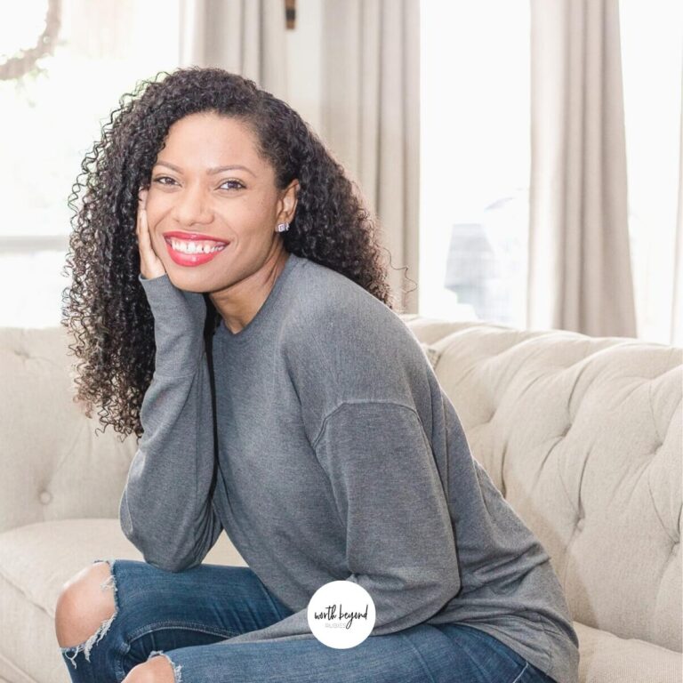 A beautiful black woman sitting on a couch smiling with her hand on her cheek and Worth Beyond Rubies logo at bottom for post on Being a Virtuous Woman and a Titus 2 Mentor