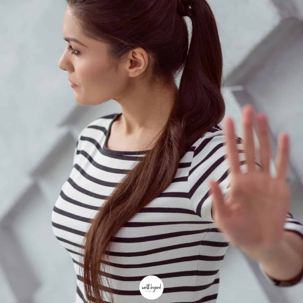 A woman in a black and white striped shirt with long brown hair in ponytail, holding her hand up in a stop motion for post on Setting Spiritual Boundaries