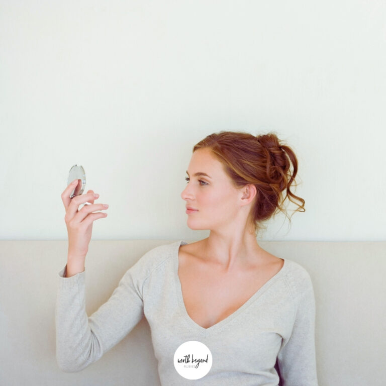 a woman in a messy bun smiling looking in a compact mirror and the Worth Beyond Rubies logo at the bottom of the image
