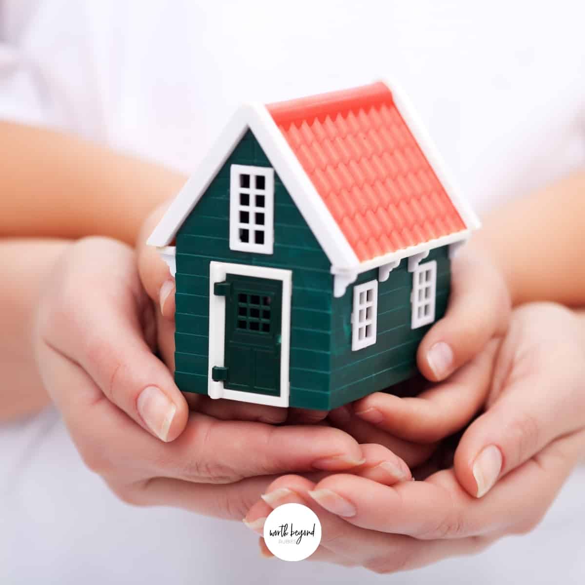 Hands holding a model of a green home and the Worth Beyond Rubies logo for post on praying over your home