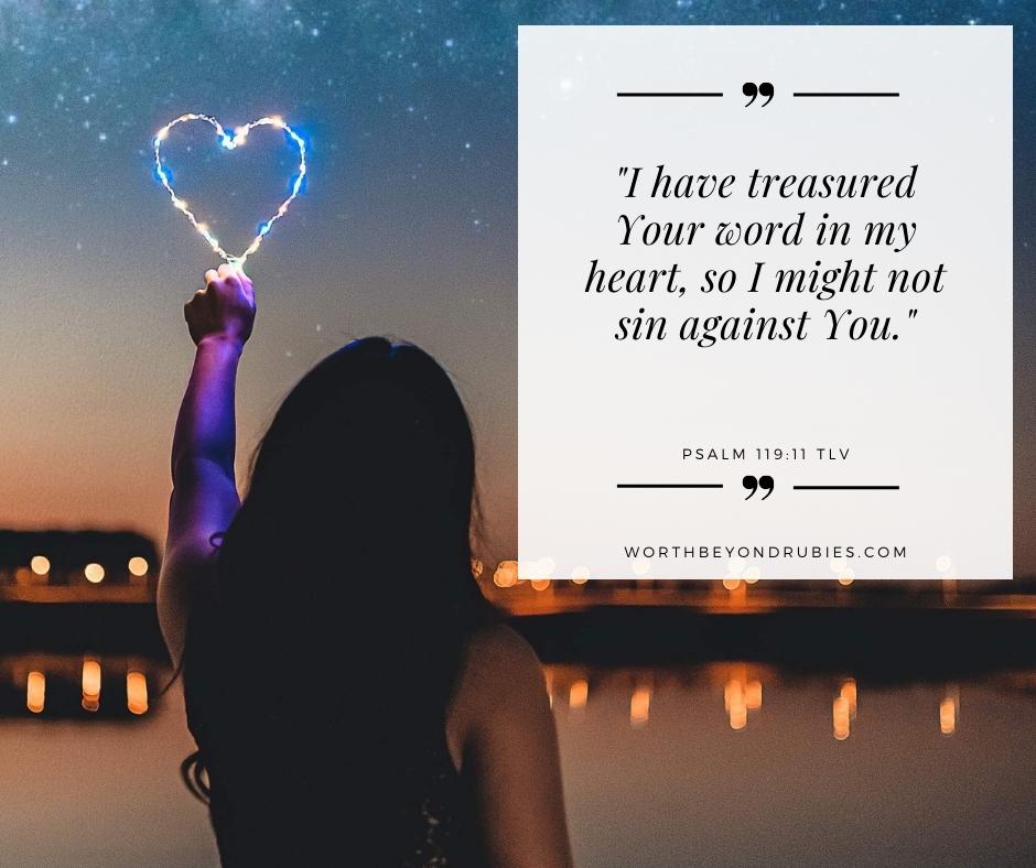 A woman standing next to water at night holding a heart shaped light and Psalm 119:11 quoted in the TLV and the link to worthbeyondrubies.com 